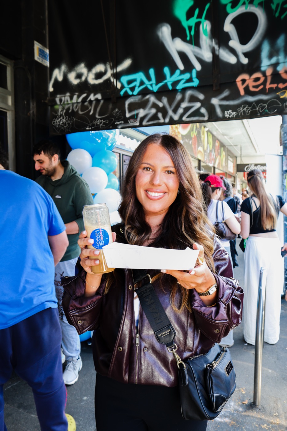 A smiling customer holding a fresh piece of bougatsa and a drink on Sydney Road